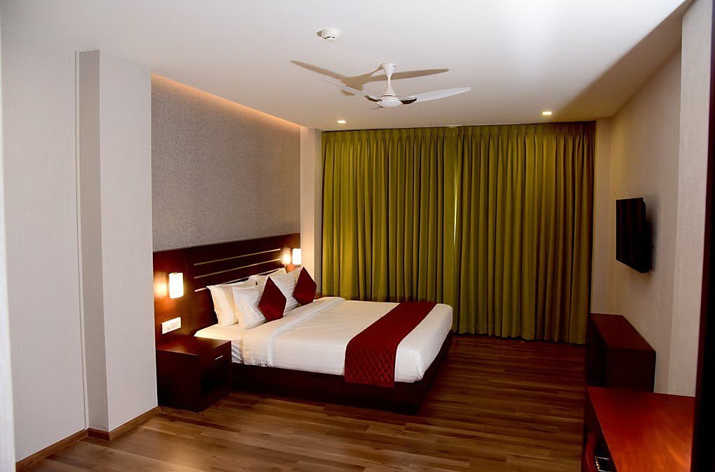 A guest room with a king-size bed in white linen and red runner, teak headboard, olive-green curtains and wooden floors